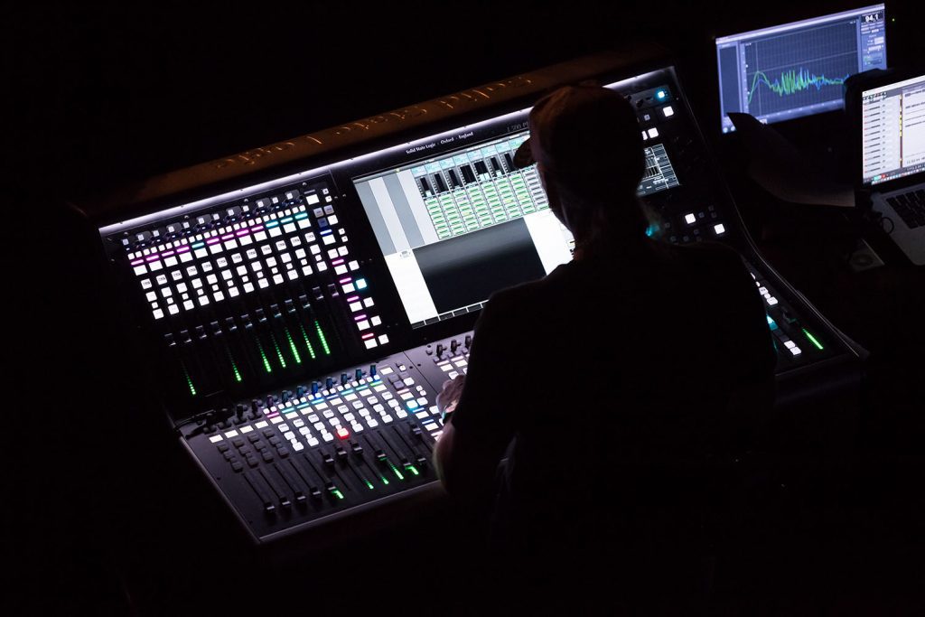 Audio engineer working on a mixing console with green lights, symbolizing our music, podcast, and voiceover services.