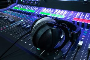 A mixing console under bluish-purple lighting with neon buttons, and headphones symbolizing our custom jingle, tag, & music services.